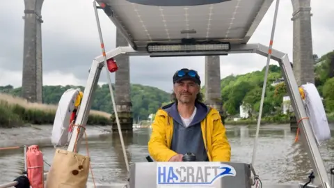 Will Mattos A man wearing a cap and a bright yellow coat. He is steering a boat in the River Tamar.