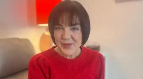 A woman with chin-length brown hair is smiling into the camera. She is wearing a red jumper.