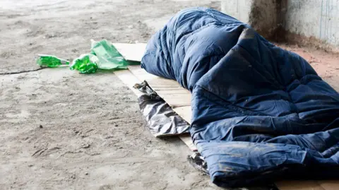 An unidentifiable person inside a blue sleeping bag, on top of a large piece of cardboard on the floor.