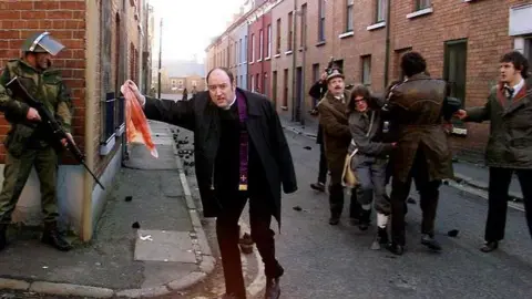 PA Media Father Edward Daly, waving a blood-stained white handkerchief surrounded by soldiers. He is wearing a black jacket and trousers, along with a white collar and a purple robe. Behind him are civilians carrying a victim and a soldier standing with camouflage clothing on holding a gun.