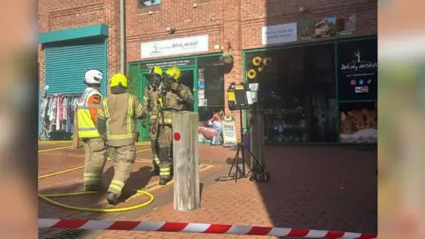 An emergency response scene outside a row of brick shopfronts on a pedestrian street. Four firefighters are in the centre of the scene, wearing full protective gear (tan fire-resistant suits, helmets, and high‑visibility markings). Fire hoses are laid out on the ground, looping across the brick‑paved walkway and leading toward the buildings. Red and white safety tape cordons off the area in the foreground.
