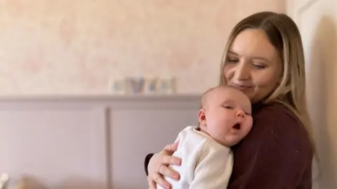 Rachel Evans holding baby Evie in their nursery at home. The behind them the wall is painted purple with the blurred name Evie on it.