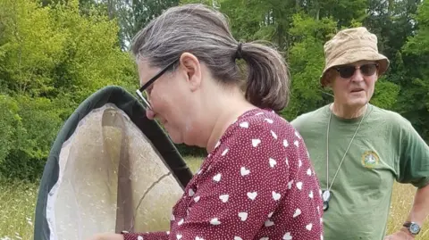 Yvonne Crouch Fiona Hutchings wears a dark red top with white hearts on it as she handles an insect inside a large net. She has her hair in a ponytail and is standing in a field. Behind her, a man wearing a green T-shirt and beige hat stands with his hands on his hips.