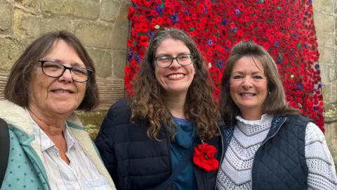 ELLEN KNIGHT/BBC Three women stood with their arms around each other in front of the poppy cascade. Steph is on the left, and is wearing a white shirt with a teal jumper. Bethan, in the middle, is wearing a blue jumper and a black winter coat, with a red knitted poppy pinned to her lapel. Trish, on the right, is wearing a white knitted jumper and a navy blue gilet. They're all smiling.
