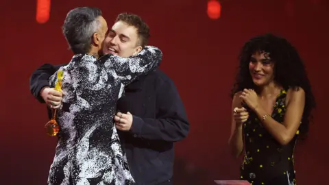 REUTERS/Isabel Infantes Robbie Williams has short grey hair and a black and white patterned formal jacket. He is embracing Sam Fender who has short brown hair and is holding a Brit Award. He is wearing a dark jacket. Olivia Dean is standing next to them with long dark hair and wearing a patterned black dress.