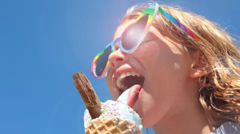 A young girl in sunglasses eating ice cream with a chocolate flake against blue sky.