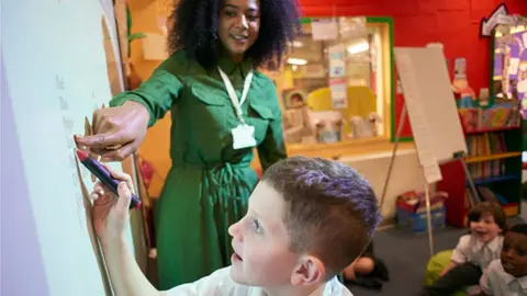 Getty Images / sturti A black teacher helps a pupil write on a whiteboard