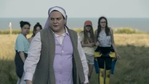 Amazon Studios Daniela Vega as Sister Maria, dressed in a nun's habit. Four young girls stand behind here and are out of focus. They appear to be standing on the grassy cliff at Bawdsey