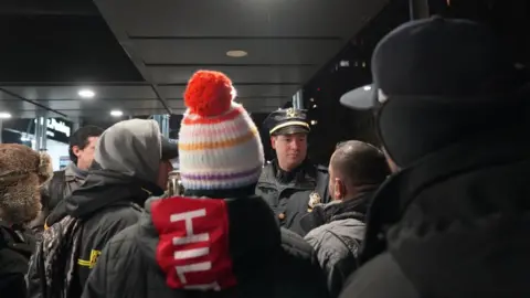 Anadolu Agency via Getty Images A police officer talks to migrants outside the Watson Hotel in New York City on 29 January, 2023