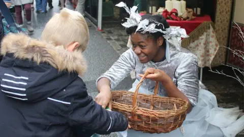 Footprints Theatre Company A woman in a white costume with a basket handing a boy something from it