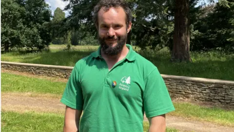 Friends of Westonbirt Arboretum Oscar Adams wearing a green Forestry England t-shirt with grass and trees around him