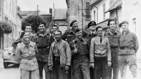Imperial War Museum 11 resistance fighters in irregular uniform, smile for the camera ahead of battle in 1944. In the background is the wing of a vintage French car and rural buildings. 