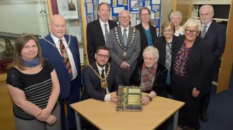 North Lanarkshire Council Several people surrounding a table, with two people sitting at the table. A woman - Helen Morrison - is sitting at the table with a tartan scarf and white hair, holding a a book with a tartan cover and titled Greetings to the women of Leningrad on the front. Sitting alongside her is a man in a suit with short dark hair, glasses and a lord provost's chain.