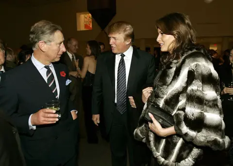 Getty Images Charles holds a drink while speaking to the Trumps. Melania wears a wool coat and clutches a purse