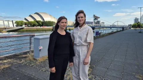Two women - Hannah Rozenberg (left) and Emer Tumilty - standing by the River Clyde and looking into the camera. The Finnieston Crane can be seen in the background. Hannah long dark hair and is wearing all black, while Emer has long dark hair and is wearing all white. It is a sunny day, with mostly blue skies. 