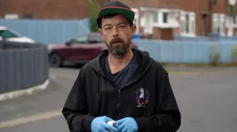 Martin Withers, who is wearing a black hooded jacket and a baseball cap, is staring directly at the camera. He is holding a can of spray paint and is wearing protective blue gloves. 