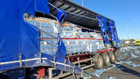 National Highways The blue side covering of a lorry is ripped, exposing stacks of plastic water bottles inside. A trail of bottles is on the road behind the lorry.