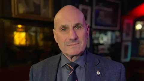 BBC/Kristian Johnson Royal Navy veteran Lt Cdr Duncan Lustig-Prean looks at the camera. He has a shaved head and is wearing a dark grey suit jacket, grey shirt, and black tie with red and white stripes. On his right lapel is a Royal Navy badge. 