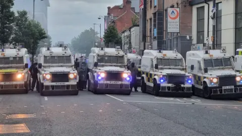 PA Media A row of police land rovers lined up across the width of a road with police in riot gear beside them