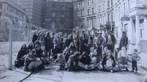Tolmers.net/Philip Thompson A black and white photo from the 1980s of a group of adults and children sitting on the road and pavement in front of a row of houses