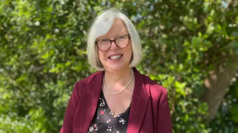 Dr Nicola Brink stands outdoors in bright sunlight. She is wearing glasses, a burgundy blazer, floral top and pearl necklace. She has short white hair and is standing in front of green trees and plants.