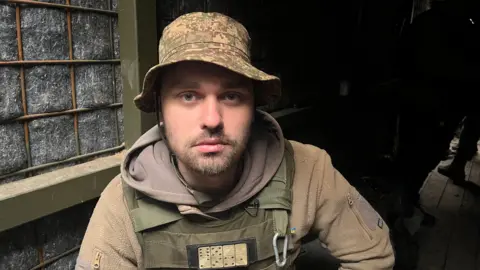 A Ukrainian soldier wears camouflage and looks directly into the camera. He has a brown hoody underneath a dark green bulletproof vest and wears a bucket hat. He has a beard and moustache and wears a forlorn expression.