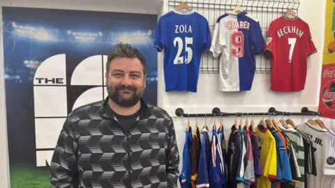 BBC A man with dark hair and a beard is wearing a black shirt with geometric white patterns. He is standing in front of two racks of brightly coloured football shirts with three on display in the upper rack.
