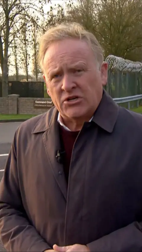 BBC journalist John Maguire stands outside RAF Fairfood. He is wearing a light jacket which has a microphone attached. Barbed wire can be seen around the entrance to the air base.
