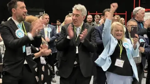 Two men and a woman punch the air while wearing light blue rosettes in front of another group of people.