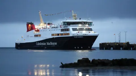 PA Media Glen Sannox di samping dermaga di bawah langit fajar biru tua. Perahu ini diterangi lampu kuning dengan garis bendera di bagian atasnya.