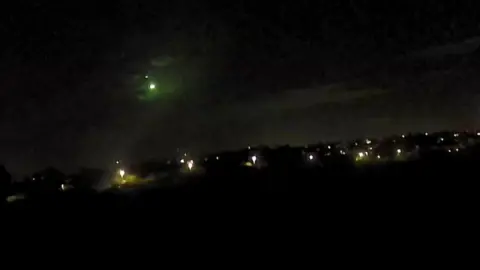 A light can be seen in the black night sky. There are street lights on the ground.