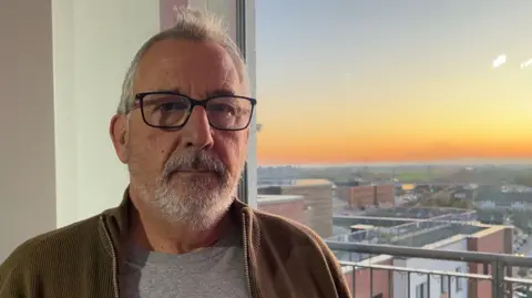 BBC News, Peterborough Michael Ferguson wears square framed glasses and has grey hair and a beard. He is inside his flat with sunset behind him outside the window.