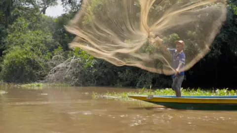 Nelayan berdiri di atas perahu kecil di Sungai Magdalena, melemparkan jaring pancing yang menyebar di udara di depannya