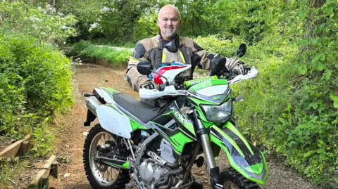 A man sits on a dirt bike on a dirt path. He is bald and he has a grey beard. He is wearing a black and brown jacket. The bike is lime green. 