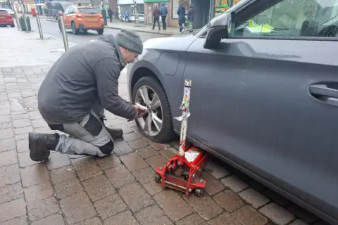 Matt Fellows A man wearing a grey top, grey hat and grey trousers removing the wheel from a grey car which has a flat tyre and is parked partly on a pavement, which is a greyish-brown colour, with a grey tarmac road in the background