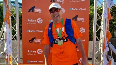 Hayden Davies at the finish line of the 2024 event. He is grinning and wearing a gold-coloured medal with a green ribbon around his neck. He is wearing an orange T-shirt which is printed with: Saffery Rotary Walk 2024, sunglasses and a cap. Behind him is a banner with Saffery Trust, Rotary in Guernsey and Saffery Rotary Walk logos and branding on it.