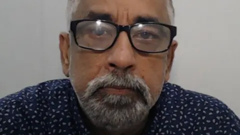 Vijay Chawla, wearing a dark blue shirt with white speckles, and with a grey beard and glasses, looks straight ahead in his police mugshot
