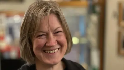 A head shot of Helen Geake smiling broadly. She has light brown bobbed hair in a long fringe brushed to her right. She is wearing a black top which can just be glimpsed.