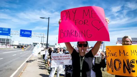 Getty Images A flight attendant pictured at a picket along a road leading to Toronto Pearson International Airport's departures hall. She is holding a hot pink sign up that reads "PAY ME FOR BOARDING" with a heart drawn on it. Next to her, a male protestor holds a bright yellow sign that says "GIVE ME MONEY". Behind her are a dozen of protestors holding various signs. One reads "Salaires de misere = pas notre Canada"