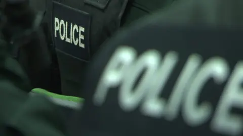 The picture shows the back of two police officers dressed in black with the word police on their jackets