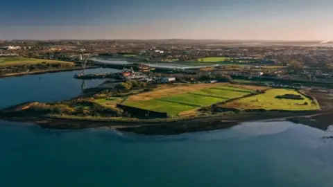 Hampshire and Isle of Wight Wildlife Trust An aerial view of the section of the peninsula Tipner West where plans are set to take place. There is a section of the island which is covered in grass field, lines by trees and surrounded by water.