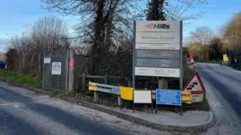 The entrance to the Lower Compton Hills Waste site in Calne, Wiltshire, with a large sign with the the Hills logo next to a road with trees either side. 