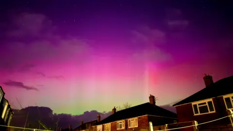 BBC Weather Watchers/Tammys Pics An evening sky with purple and pink hues and stars can be seen faintly at the top of the photo. Green hues can be seen in the far distance in between houses. 