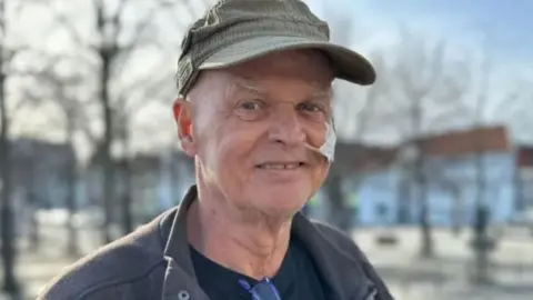 Kian Reme smiles at the camera. He is wearing a green hat and has a tube coming out of his nose. He is wearing a navy jacket and is standing in front of some buildings and trees