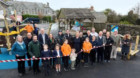 Environment Agency A group of adults and children standing behind a red and white plastic tape. Behind them is the playground and school building. It is a grey day.