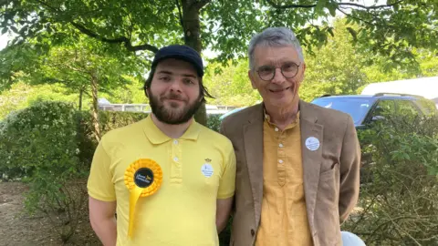 Emma Howgego/BBC On the left is Ariel Cahn, who wears a yellow t-shirt, baseball cap and a yellow Liberal Democrat rosette. On the right is Martin Cahn, who wears glasses, a light-brown jacket and a golden-coloured shirt. 