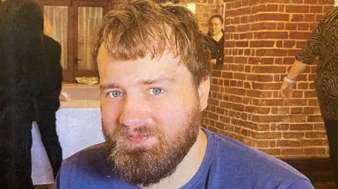 Tom James is looking directly at the camera in a close-up photograph. He is indoors. Behind him is a bare brick wall and several people, and two tables with white tablecloths on.