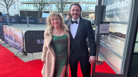 Gabby Mason and Leyton Hunnissett are smiling for a photo on a red carpet outside Cineworld in Jersey, at the Bergerac premiere. They are wearing black tie.