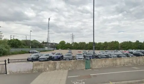 Google The car park seen from the road, with about 40 cars parked in it in daylight. There are trees lining one edge and a metal fence along the left border, and the overhead wires and gantries of the train line.