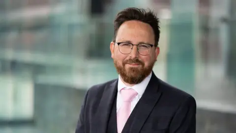 Hefin David outside the Senedd, he is wearing a grey suit and a pink tie, with glasses and a full beard